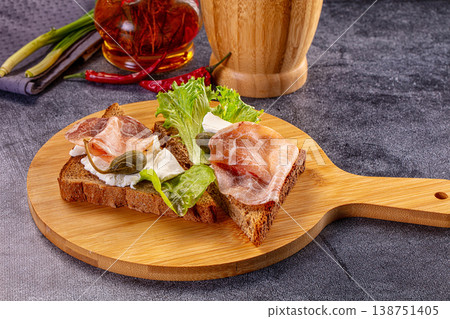 Gourmet rye bread toast with cured meat, soft cheese, and capers on a wooden board. 138751405