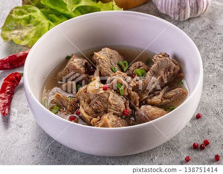 Savory meat and bone broth soup in a white bowl with garlic, chili, and herbs on a rustic grey table 138751414