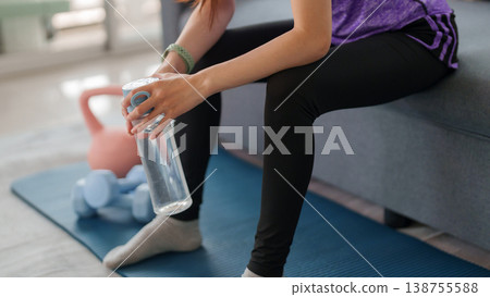 Asian woman hydrating home workout taking break after exercise session sitting on sofa holding water bottle practicing fitness wellness lifestyle at home Asian woman hydrating home workout taking break after exercise session sitting on sofa holding water bottle practicing fitness wellness lifestyle at home 138755588