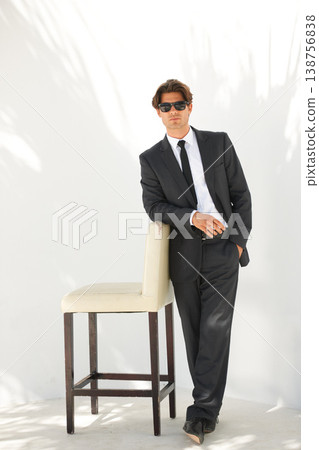Portrait, fashion and a business man with a chair in studio on a white background for corporate style. Handsome, sunglasses and formal with a confident young professional employee leaning on a seat 138756838