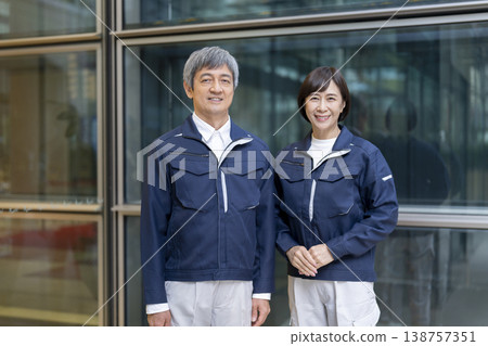 A man and woman in their 60s, wearing work clothes and smiling at the camera; they appear to be apartment building managers and facility staff. (Image) A man and woman in their 60s, wearing work clothes and smiling at the camera; they appear to be apartment building managers and facility staff. (Image) 138757351