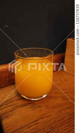 Glass of orange juice with ice cubes placed on a wooden table against a dark wall, showcasing a refreshing beverage in a casual setting Glass of orange juice with ice cubes placed on a wooden table against a dark wall, showcasing a refreshing beverage in a casual setting 138757639