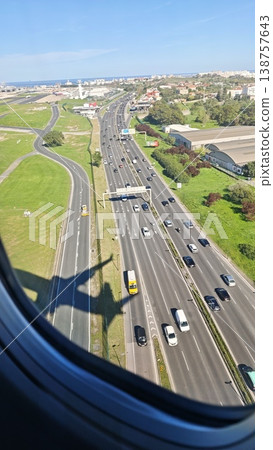 Aerial view of a busy highway with vehicles traveling, green fields and buildings visible, captured from an airplane window during descent near a coastal city Aerial view of a busy highway with vehicles traveling, green fields and buildings visible, captured from an airplane window during descent near a coastal city 138757643