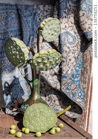 Several green lotus seed pods rise from a slender bronze-colored vase, their unique, honeycomb textures catching the light. Below, a single large pod and a handful of round seeds are strewn across a Several green lotus seed pods rise from a slender bronze-colored vase, their unique, honeycomb textures catching the light. Below, a single large pod and a handful of round seeds are strewn across a 138758251