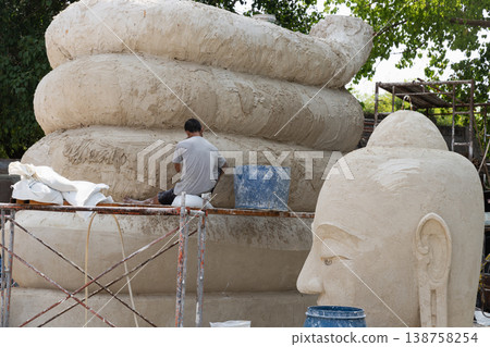 A sculptor sits on a metal scaffolding, working on the massive, coiled base of a monumental Buddha statue. In the foreground, a large, finished sculpted head with serene facial features and elongated A sculptor sits on a metal scaffolding, working on the massive, coiled base of a monumental Buddha statue. In the foreground, a large, finished sculpted head with serene facial features and elongated 138758254