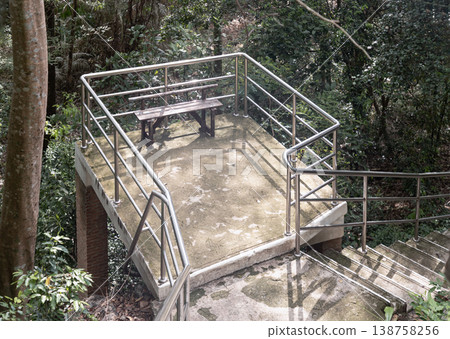 A small, elevated concrete platform with a single wooden bench overlooks a dense, green forest. Modern stainless steel railings enclose the overlook for safety, connecting to a set of stone or A small, elevated concrete platform with a single wooden bench overlooks a dense, green forest. Modern stainless steel railings enclose the overlook for safety, connecting to a set of stone or 138758256