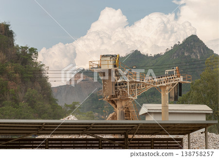 A large-scale industrial conveyor prominently anchors the scene, highlighting the scale of operations at this quarry. Beyond the machinery, the landscape transitions into rugged mountains that rise 138758257