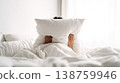 Person lies in bed hiding face behind pillow in bright bedroom for privacy and comfort 138759946