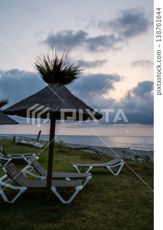 在地中海的法國科西嘉島,黎明時分,茅草屋頂的遮陽傘和沙灘床沿著海岸線排列。 在地中海的法國科西嘉島,黎明時分,茅草屋頂的遮陽傘和沙灘床沿著海岸線排列。 138761644