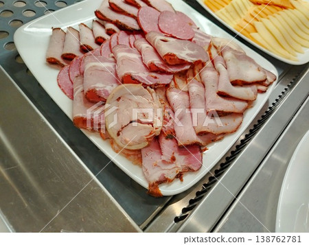 Assorted cold cuts including sliced ham and salami arranged on a white platter, with cheese slices visible in the background on a stainless steel surface 138762781
