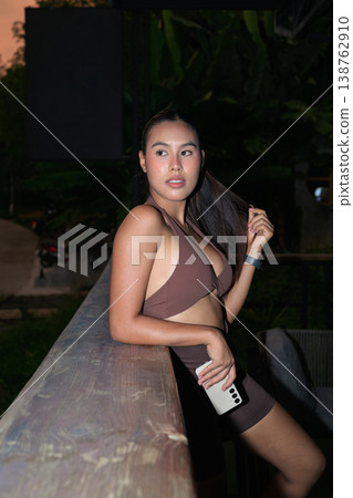 Young Thai Fitness Influencer Holding Smartphone on Balcony at Dusk Modern Digital Lifestyle Concept 138762910
