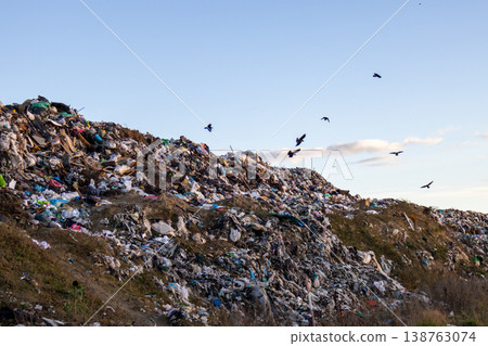 A vast heap of trash sprawls against the horizon while birds glide overhead, creating a stark contrast with the peaceful twilight 138763074
