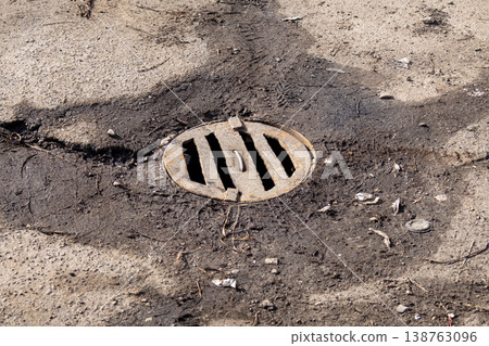 A storm drain sits in the center of a cracked concrete surface, surrounded by dirt and litter on a bright day, showing signs of wear and neglect A storm drain sits in the center of a cracked concrete surface, surrounded by dirt and litter on a bright day, showing signs of wear and neglect 138763096