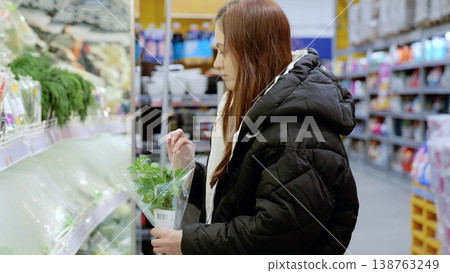 Young woman selecting fresh parsley in the produce section of a grocery store, examining the quality and freshness of the herbs while shopping for ingredients Young woman selecting fresh parsley in the produce section of a grocery store, examining the quality and freshness of the herbs while shopping for ingredients 138763249