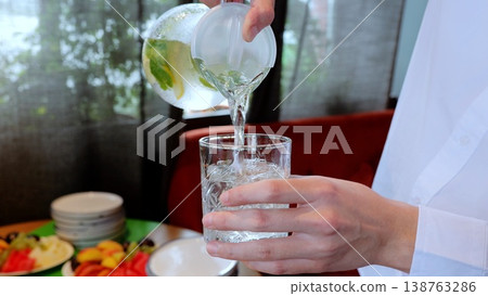 Waiter pouring chilled water with lemon and mint into glass, fruit platter decorating restaurant background 138763286