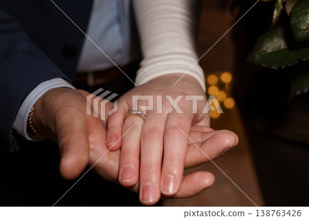 Close-up of engaged couple holding hands with diamond ring on woman's finger, man in suit, romantic lighting Close-up of engaged couple holding hands with diamond ring on woman's finger, man in suit, romantic lighting 138763426