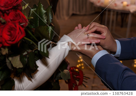 Groom placing wedding ring on bride finger holding red roses bouquet during romantic ceremony Groom placing wedding ring on bride finger holding red roses bouquet during romantic ceremony 138763428