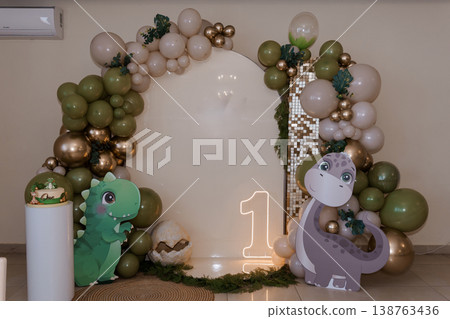 First birthday dinosaur party backdrop with green and gold balloons, neon number one sign, cartoon dino cutouts, and cake on white pedestal. First birthday dinosaur party backdrop with green and gold balloons, neon number one sign, cartoon dino cutouts, and cake on white pedestal. 138763436