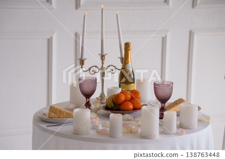 Elegant round table setting with champagne bottle, purple goblets, oranges, cheese, and lit candles on white tablecloth against classic white wall paneling. 138763448