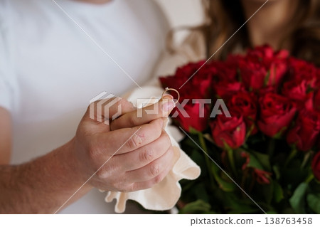 Close-up of male hand holding diamond engagement ring over red roses with female partner in background for romantic proposal scene. Close-up of male hand holding diamond engagement ring over red roses with female partner in background for romantic proposal scene. 138763458