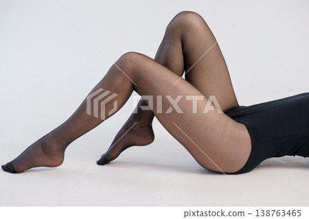 Close up of woman legs in black sheer tights lying on white floor in minimalist studio setting. Close up of woman legs in black sheer tights lying on white floor in minimalist studio setting. 138763465