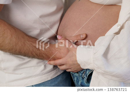 Pregnant woman and partner holding hands on baby bump in white shirts and jeans 138763721