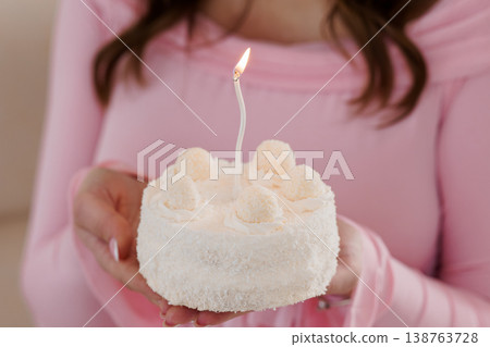 Young woman holding small white birthday cake with single lit candle and pink clothing closeup Young woman holding small white birthday cake with single lit candle and pink clothing closeup 138763728