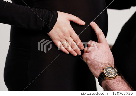 Pregnant couple holding baby bump with wedding rings and luxury watch on black background Pregnant couple holding baby bump with wedding rings and luxury watch on black background 138763730