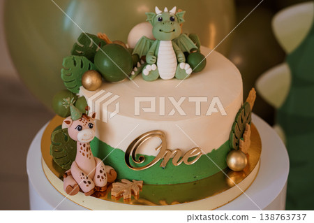 First birthday cake decorated with green dragon and giraffe figurines on gold board with monstera leaves and balloons. First birthday cake decorated with green dragon and giraffe figurines on gold board with monstera leaves and balloons. 138763737