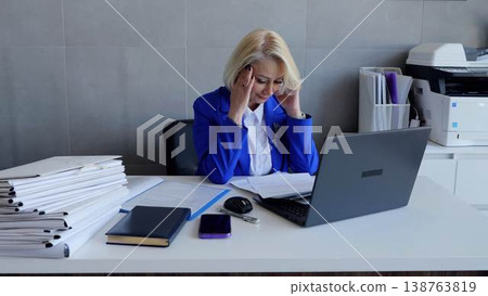 Overwhelmed senior businesswoman with a headache working on a laptop in a busy office surrounded by paperwork 138763819