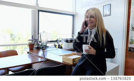 Professional female executive confidently manages multiple tasks in her contemporary office, conversing on the phone and savoring a cup of coffee Professional female executive confidently manages multiple tasks in her contemporary office, conversing on the phone and savoring a cup of coffee 138763820
