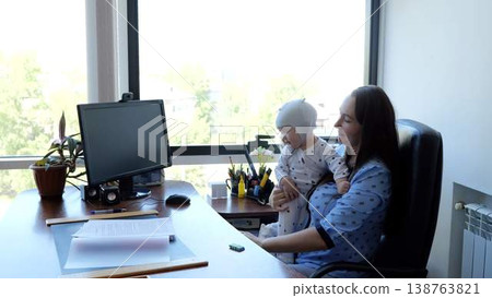 Businesswoman sitting at her desk, holding her baby in her lap while working on her computer in her home office, demonstrating work life balance. Businesswoman sitting at her desk, holding her baby in her lap while working on her computer in her home office, demonstrating work life balance. 138763821