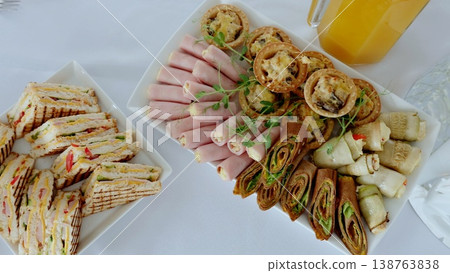 Catering service displaying gourmet finger foods ham cheese rolls, club sandwiches, mini quiches, stuffed pancakes, fresh juice arranged on crisp white linen 138763838