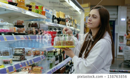 Young woman shopping for dessert, holding a packaged cake and looking at other options in the refrigerated bakery section of a grocery store 138763856