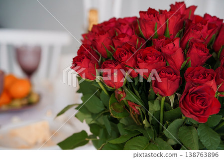 Large bouquet of vibrant red roses with green leaves on a blurred dinner table setting with wine glass and fruit in background. Large bouquet of vibrant red roses with green leaves on a blurred dinner table setting with wine glass and fruit in background. 138763896