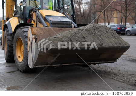 Yellow excavator loader bucket filled with dark soil on a city street construction site. Yellow excavator loader bucket filled with dark soil on a city street construction site. 138763929