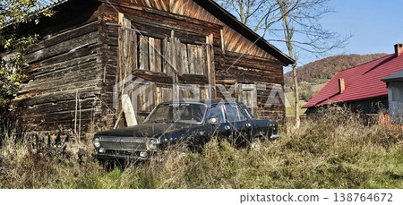 Abandoned vintage car sits overgrown in tall grass beside a weathered wooden barn in a rural countryside. 138764672