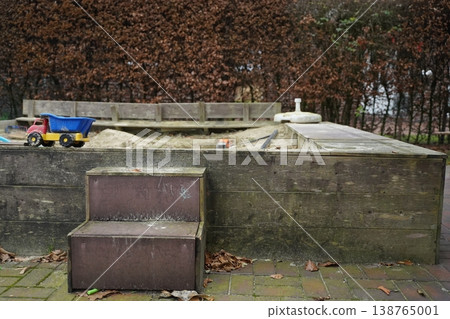 Wooden sandbox with a blue toy dump truck and a bench on a leaf-covered patio beside a low wall and trees. Quiet playground scene. Wooden sandbox with a blue toy dump truck and a bench on a leaf-covered patio beside a low wall and trees. Quiet playground scene. 138765001