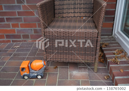 Wicker armchair on brick patio beside glass door, with toy cement mixer truck in front and dry leaves scattered. Outdoor still life with furniture and toy. 138765002
