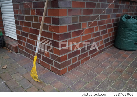 Yellow plastic rake leaning against brick wall on a paved patio; a green bag sits nearby. Outdoor corner scene with yard tool. Simple outdoor maintenance moment. Yellow plastic rake leaning against brick wall on a paved patio; a green bag sits nearby. Outdoor corner scene with yard tool. Simple outdoor maintenance moment. 138765005