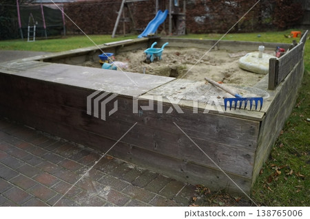 Wooden sandpit in a yard with toys, a blue rake, and a small slide in the background. Outdoor play area for children. Wooden sandpit in a yard with toys, a blue rake, and a small slide in the background. Outdoor play area for children. 138765006