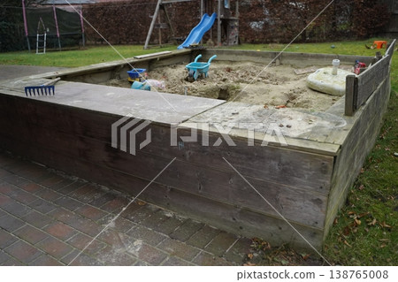Wooden sandbox in a yard with sand, toy wheelbarrow, rake, and blue slide in the background. Outdoor play area for children. 138765008
