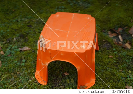 Orange plastic stool turned upside down on mossy ground with a small patch of mud on one side. Outdoor scene focuses on a worn seat. Orange plastic stool turned upside down on mossy ground with a small patch of mud on one side. Outdoor scene focuses on a worn seat. 138765018