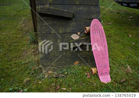 Pink skateboard leaning against weathered wooden crate on grassy ground outdoors, with scattered leaves. A casual skate scene in a park-like setting. 138765019