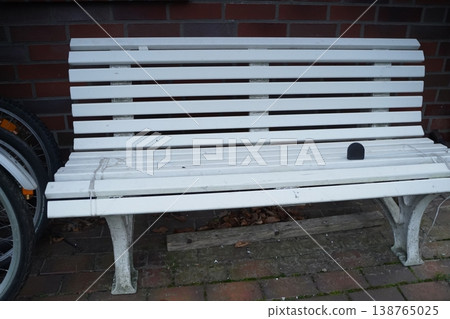 White wooden bench with scuffs in front of a dark brick wall; part of a bicycle wheel at left. Outdoor seating by a wall. 138765025