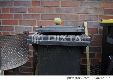 Outdoor trash bin with open lid against a brick wall; a tennis ball sits on top, with a wooden handle and metal basket nearby. The scene shows discarded items by a wall. Outdoor trash bin with open lid against a brick wall; a tennis ball sits on top, with a wooden handle and metal basket nearby. The scene shows discarded items by a wall. 138765026