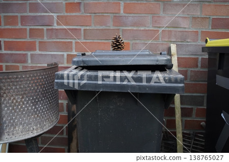 A dark plastic trash bin with an open lid stands against a brick wall, with a pine cone on top and tools nearby. Everyday outdoor waste area scene. A dark plastic trash bin with an open lid stands against a brick wall, with a pine cone on top and tools nearby. Everyday outdoor waste area scene. 138765027