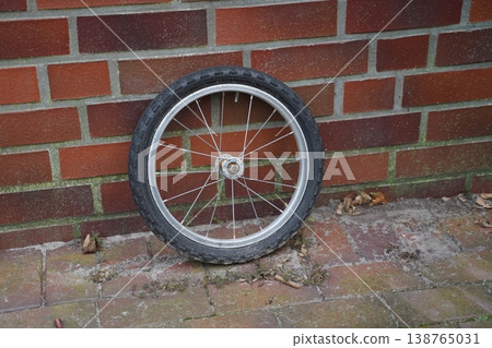 Single bicycle wheel leaning against a brick wall on a paved surface with scattered leaves. A lone wheel rests by a wall outdoors. Single bicycle wheel leaning against a brick wall on a paved surface with scattered leaves. A lone wheel rests by a wall outdoors. 138765031