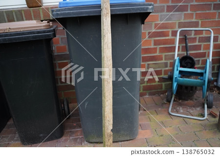 Two large dark trash bin stand on a brick patio by a brick wall; a wooden post in front and a blue hose reel at right. Outdoor storage area scene. Two large dark trash bin stand on a brick patio by a brick wall; a wooden post in front and a blue hose reel at right. Outdoor storage area scene. 138765032