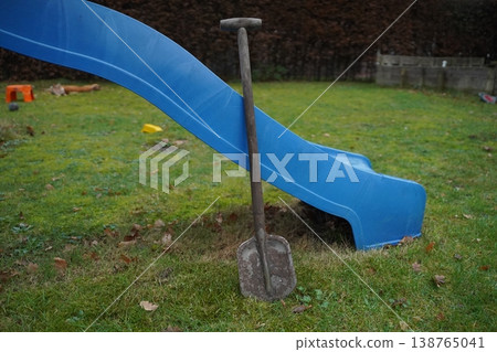 Metal shovel standing in grass beside a blue playground slide on a yard. Outdoor play area scene. 138765041